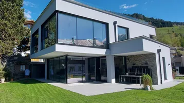 Moderne Villa mit großen Glasfronten, gepflegtem Rasen und einer sonnigen Terrasse.