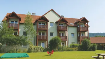 Mehrfamilienhaus mit hellgrüner Fassade, Holzbalkonen und einem gepflegten Gartenbereich mit Spielgeräten.