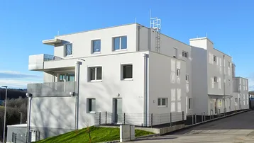 Modernes Mehrfamilienhaus mit weißer Fassade und Balkonen in grüner Umgebung.