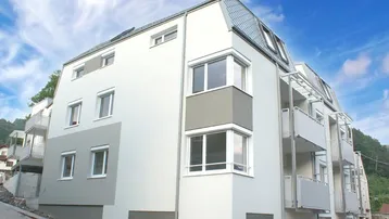 Modernes Mehrfamilienhaus mit hellgrauer Fassade, großen Fenstern und Balkonen in grüner Umgebung.