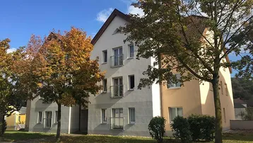 Mehrfamilienhaus mit heller Fassade, kleinen Balkonen und umgebenden Bäumen in herbstlicher Färbung.
