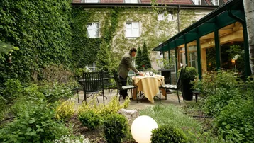 Ein Mann deckt einen Tisch in einem grünen Gartenbereich mit üppiger Vegetation und einem historischen Gebäude im Hintergrund.