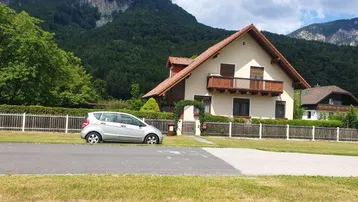 Traditionelles Haus mit Satteldach und Balkon, umgeben von grünen Bergen und einem gepflegten Vorgarten.