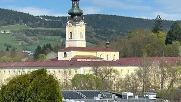 Panoramablick auf ein historisches Klostergebäude mit Kirchturm inmitten einer grünen Hügellandschaft.