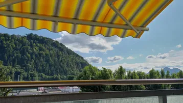 Blick vom Balkon mit gelber Markise auf bewaldete Berge und weite Landschaft.