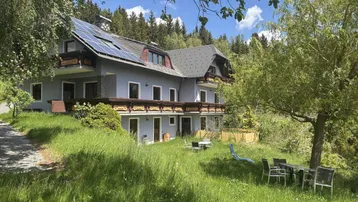 Haus mit Solaranlage, Balkonen und Gartenmöbeln in einer grünen Umgebung mit Bergblick.