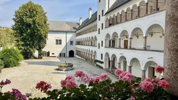 Historischer Innenhof eines Anwesens mit Arkaden, weißen Wänden und blühenden Geranien im Vordergrund.