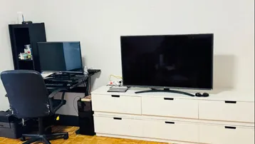 Wohnbereich mit Fernseher auf einem Sideboard und einem Arbeitsplatz mit Schreibtisch und Bürostuhl.