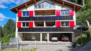 Modernes Mehrfamilienhaus mit roten Fensterläden, Solarpaneelen und Bergblick in grüner Umgebung.