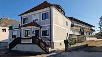 Graues Gebäude mit braunem Dach und Holzgeländer an der Eingangstreppe unter blauem Himmel.