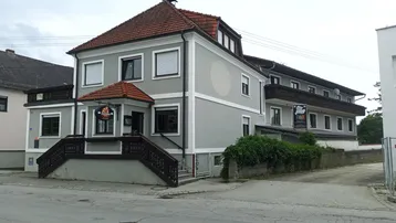 Graues Gebäude mit rotem Ziegeldach, Rollläden und einem Eingangsbereich mit Treppe.
