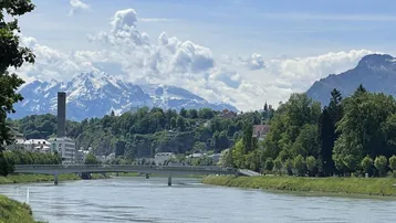 Panoramablick auf einen Fluss mit Brücke, Stadt am Ufer und schneebedeckten Bergen im Hintergrund unter blauem Himmel.