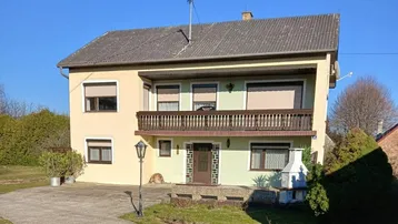 Zweistöckiges Haus mit Balkon, gepflasterter Auffahrt und Satteldach unter blauem Himmel.