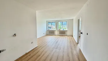 Helles, leeres Wohnzimmer mit neuem Laminatboden und Blick ins Grüne durch die Fenster.