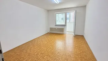 Helles, leeres Zimmer mit Parkettboden und Zugang zu einem kleinen Balkon.