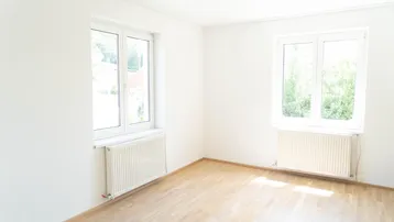 Helles, leeres Zimmer mit zwei Fenstern, Parkettboden und Heizkörpern, bereit zur individuellen Gestaltung.