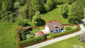 Luftaufnahme eines Einfamilienhauses mit Grundstücksumrandung in grüner Hügellandschaft.