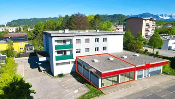 Luftaufnahme eines Gewerbegebäudes mit angrenzendem Parkplatz und Wohnhaus, umgeben von grüner Landschaft und Bergen.