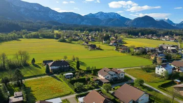 Weitläufige Landschaft mit Wohnsiedlung, grünen Feldern und majestätischen Bergen im Hintergrund.