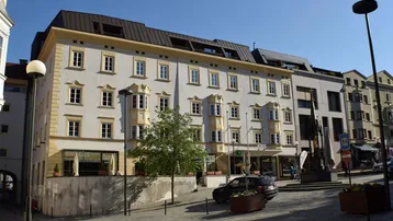 Historisches Gebäude mit heller Fassade, vielen Fenstern und Geschäften im Erdgeschoss an einer belebten Straße.