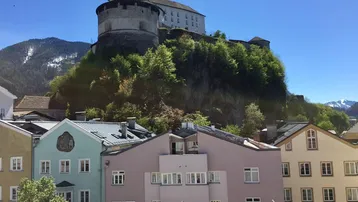 Historische Burg auf einem Hügel mit bunten Häusern im Vordergrund und Bergblick.