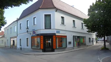Eckgebäude mit hellblauer Fassade und rotem Ziegeldach, im Erdgeschoss befindet sich ein Geschäft.