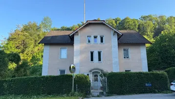 Klassisches Gebäude mit hellbeiger Fassade, umgeben von grünen Hecken und Bäumen.
