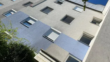 Fassade eines Mehrfamilienhauses mit verschiedenen Fenstergrößen und Rollläden unter blauem Himmel.