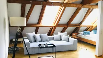 Helles Dachgeschoss-Wohnzimmer mit sichtbaren Holzbalken, modernem Sofa und Schlafbereich im Hintergrund.