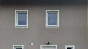 Moderne Hausfassade in Grau mit weißen Fenstern und Eingangstür unter einem Satteldach.