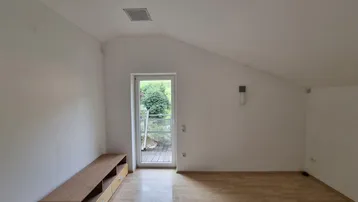 Helles Zimmer mit Holzboden, Dachschräge und Zugang zu einem kleinen Balkon oder einer Terrasse.