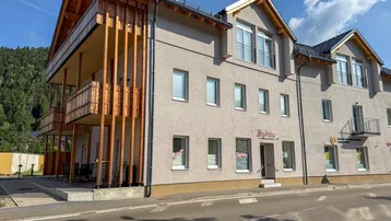Modernes Gebäude mit grauer Fassade, Holzbalkonen und Blick auf grüne Berge unter blauem Himmel.