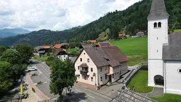 Luftaufnahme eines malerischen Dorfes mit Kirche, Häusern und grünen Hügeln unter blauem Himmel.