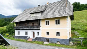 Traditionelles Bauernhaus mit Holzbalkon und Schindeldach in ländlicher Umgebung, umgeben von grünen Hügeln.