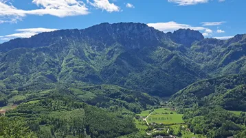 Weitläufige Berglandschaft mit grünen Wäldern, einem Tal und einem kleinen Dorf unter blauem Himmel.