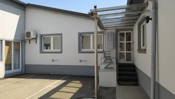 Eingangsbereich eines Gebäudes mit Vordach, Klimaanlage und Parkplätzen für Gäste.