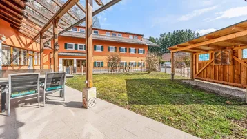 Große gepflasterte Terrasse mit Holzpergola und Gartenmöbeln, Blick auf das Hauptgebäude und einen Carport.