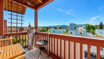 Sonniger Balkon mit Holztisch und Stühlen, gefliestem Boden und weitem Blick über die grüne Landschaft und Berge.