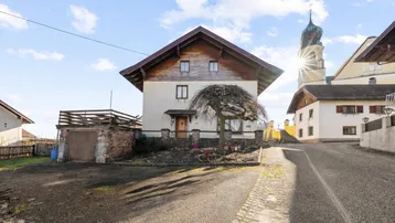 Traditionelles Einfamilienhaus mit Satteldach und angrenzender Garage in ländlicher Umgebung mit Kirchturm im Hintergrund.