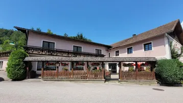 Traditionelles Gebäude mit überdachter Terrasse und Holzbalkonen, umgeben von grüner Landschaft und blauem Himmel.