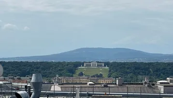 Fernblick auf das Schloss Schönbrunn und die Gloriette, umgeben von grünen Wäldern und Bergen.