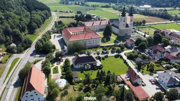 Luftaufnahme eines Dorfes mit einem großen Klosterkomplex und umliegender ländlicher Landschaft.
