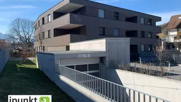 Modernes Mehrfamilienhaus mit dunkler Holzfassade und Zufahrt zur Tiefgarage unter blauem Himmel.