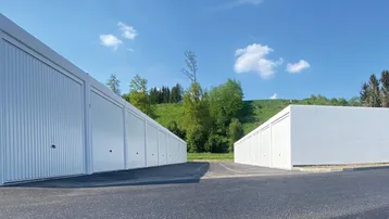 Reihen von weißen Garagenboxen mit Asphaltvorplatz vor einer grünen Hügellandschaft unter blauem Himmel.