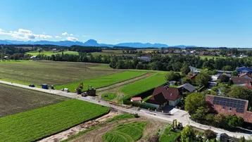Luftaufnahme einer ländlichen Gegend mit Feldern, Häusern und Bergen im Hintergrund unter blauem Himmel.