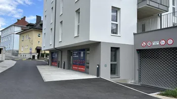 Modernes Gebäude mit hellgrauer Fassade, Tiefgarageneinfahrt und Geschäftsflächen im Erdgeschoss.