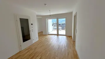 Helles, leeres Wohnzimmer mit Parkettboden und Zugang zu einem Balkon mit Bergblick.