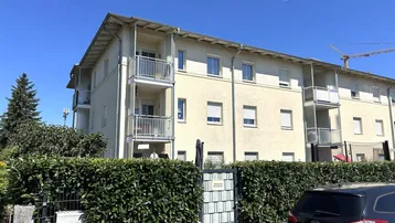 Mehrfamilienhaus mit mehreren Balkonen und gepflegter Hecke im Vordergrund unter blauem Himmel.