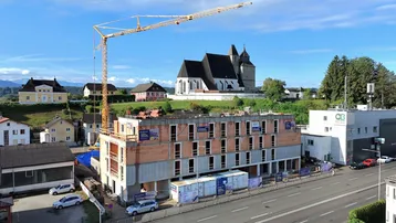 Luftaufnahme eines im Bau befindlichen modernen Wohngebäudes mit Kran und Kirche im Hintergrund.