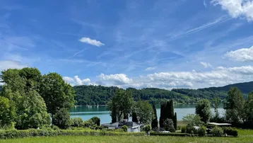 Panoramablick auf einen malerischen See mit grünen Hügeln und einem Haus im Vordergrund unter blauem Himmel.